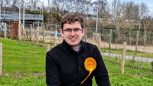 Grant Toghill sitting on a wall wearing a Scottish Lib Dem rosette.