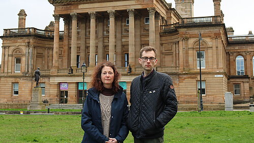 Anne Hannigan and Jack Clark standing next to each other, looking unhappy with Paisley Town Hall in the background.