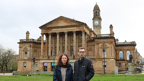 Anne Hannigan and Jack Clark standing next to each other, looking unhappy with Paisley Town Hall in the background.